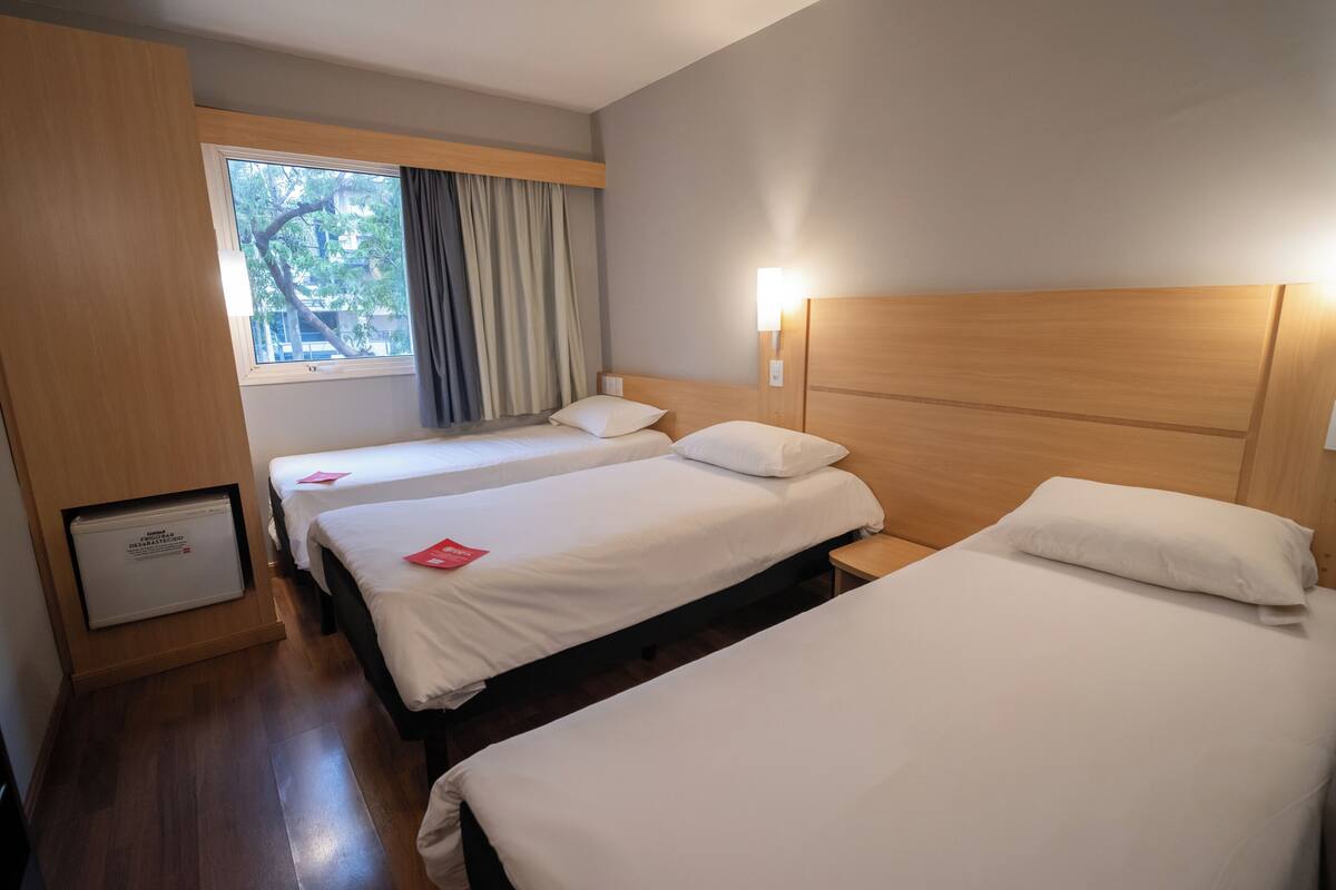 A hotel room with two beds, a window with curtains, a wall-mounted light, and a small cabinet.