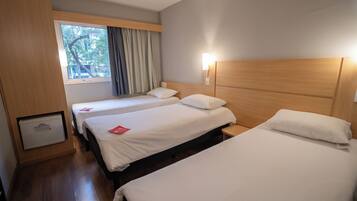 Ein Hotelzimmer mit zwei Betten, einem Fenster mit Vorhängen, einer Wandlampe und einem kleinen Schrank.