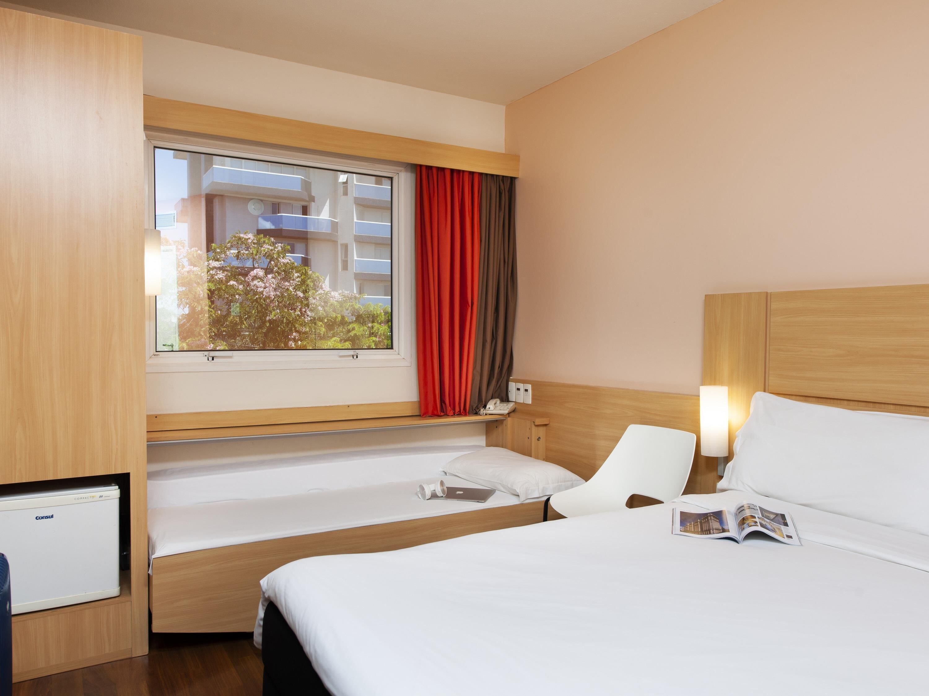 A hotel room with a bed, a desk, a chair, a window with curtains, and a view of buildings and trees.