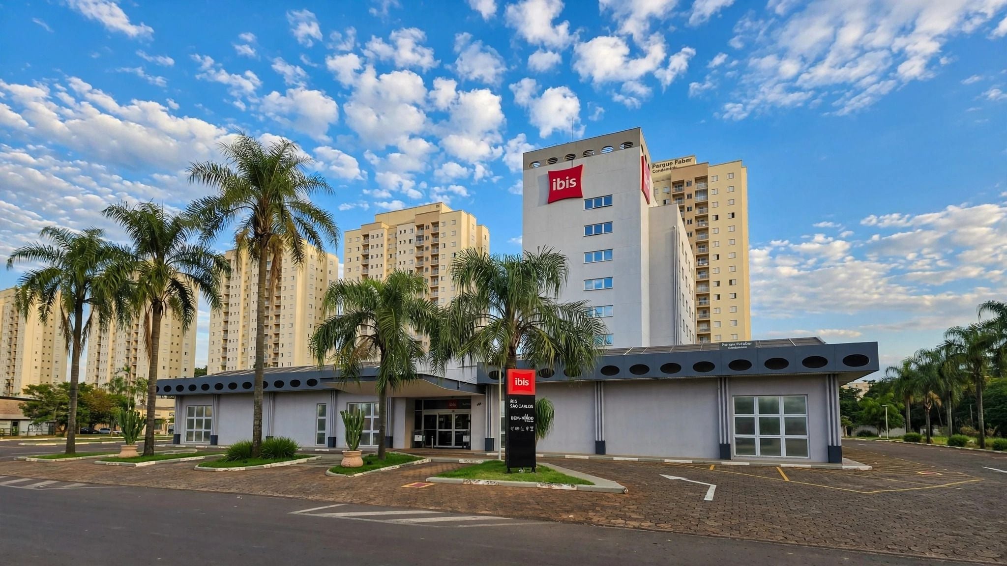 A modern hotel with a prominent ibis logo, surrounded by palm trees and adjacent high-rise buildings.