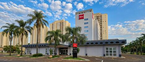 Un hotel moderno con un llamativo logotipo de ibis, rodeado de palmeras y edificios altos contiguos.