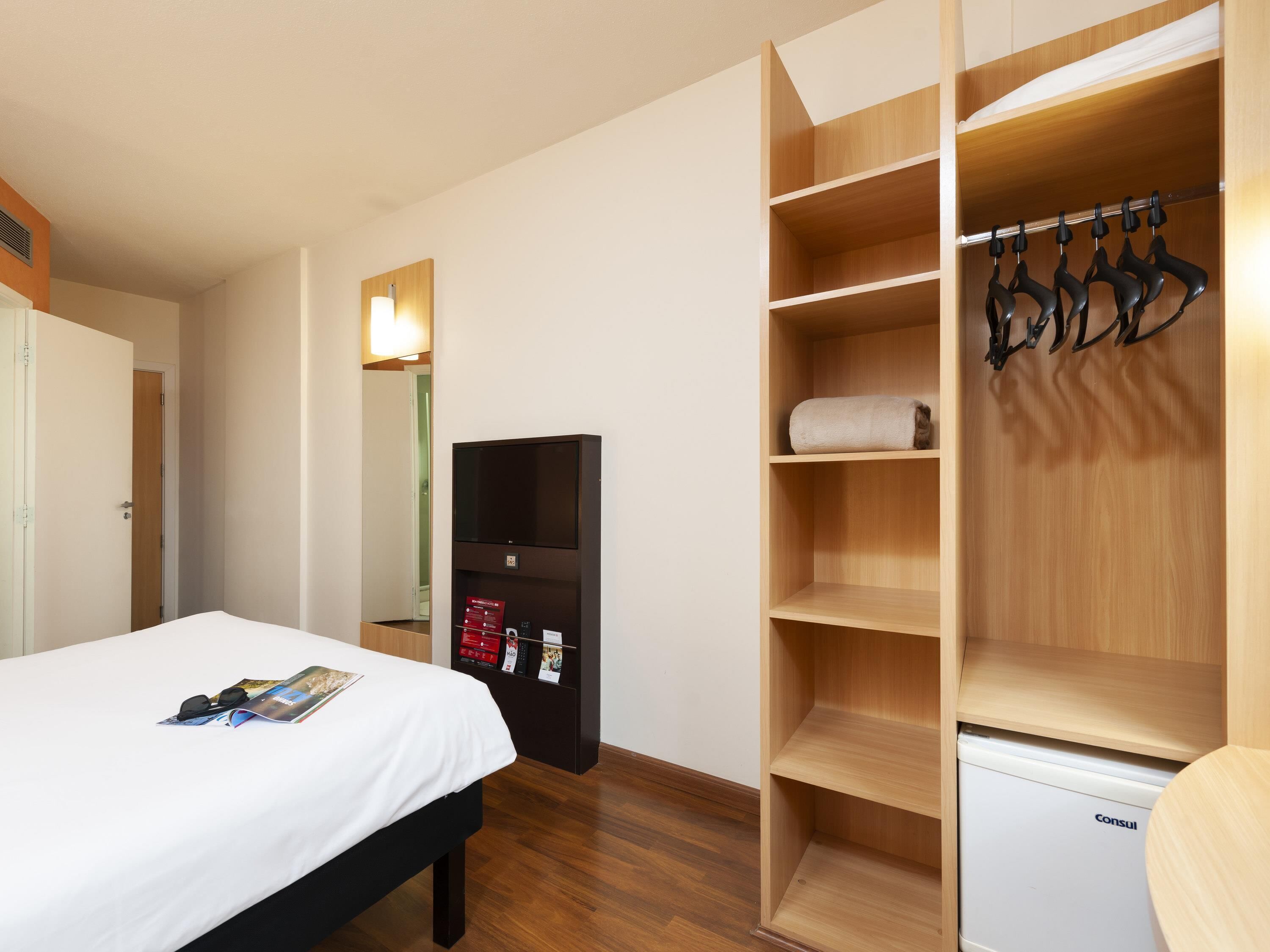 A hotel room with a bed, a wardrobe with hanging space, a small desk with a lamp, and a white bedside table.