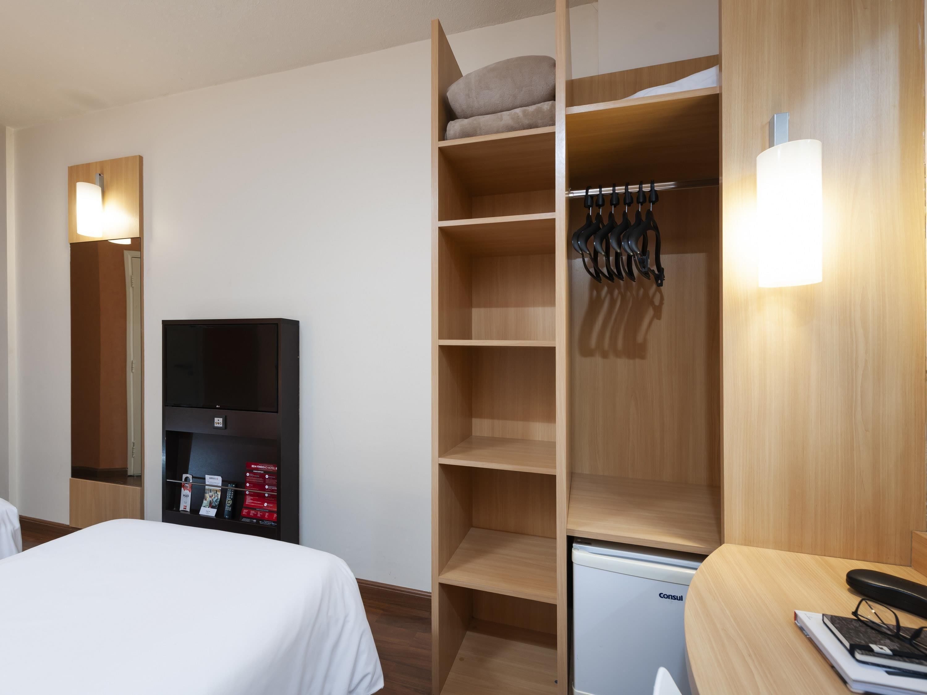 A hotel room with a bed, a TV, a minibar, a wardrobe, and a desk with a lamp.