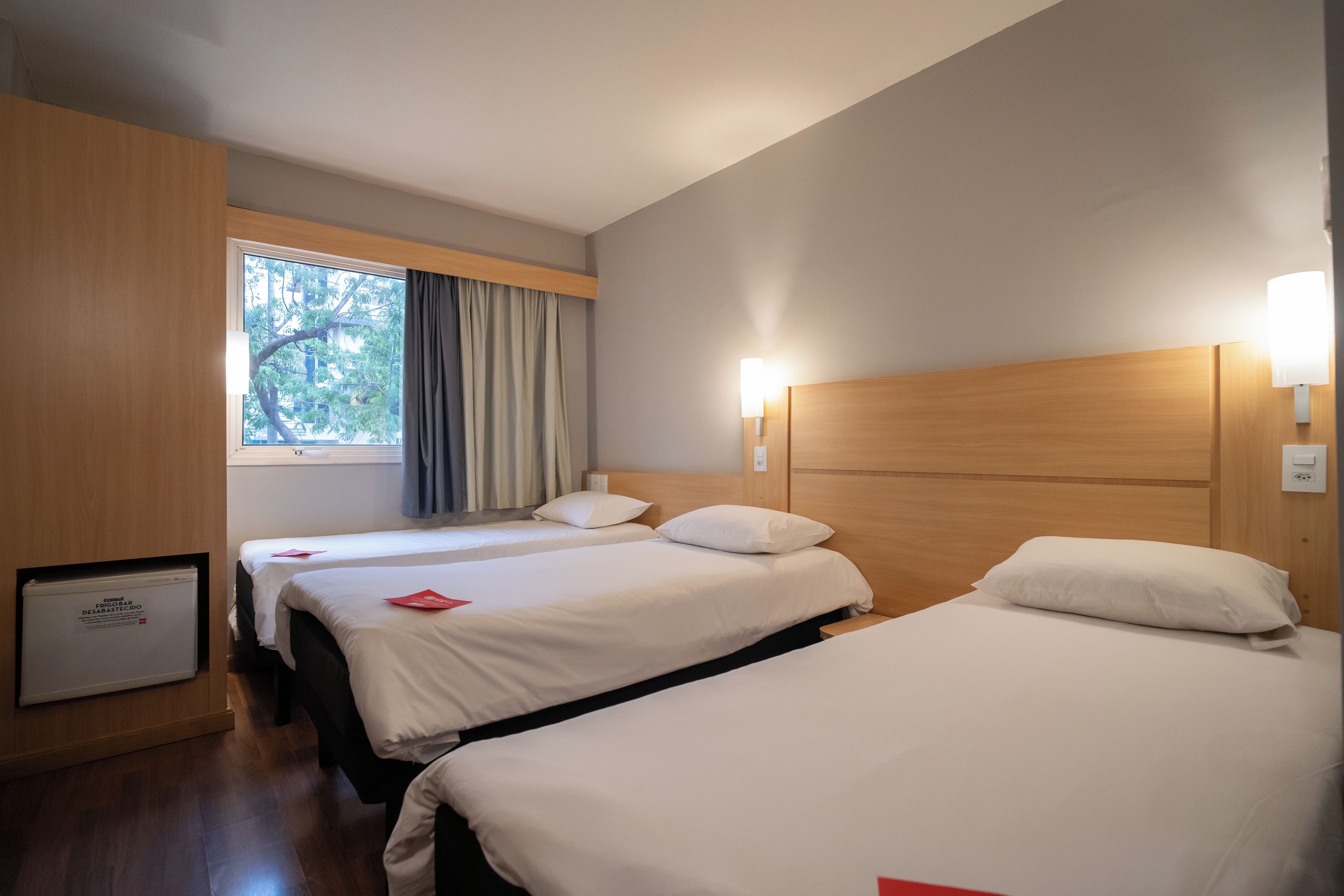 A hotel room with three beds, a window with curtains, and a wall-mounted light fixture.