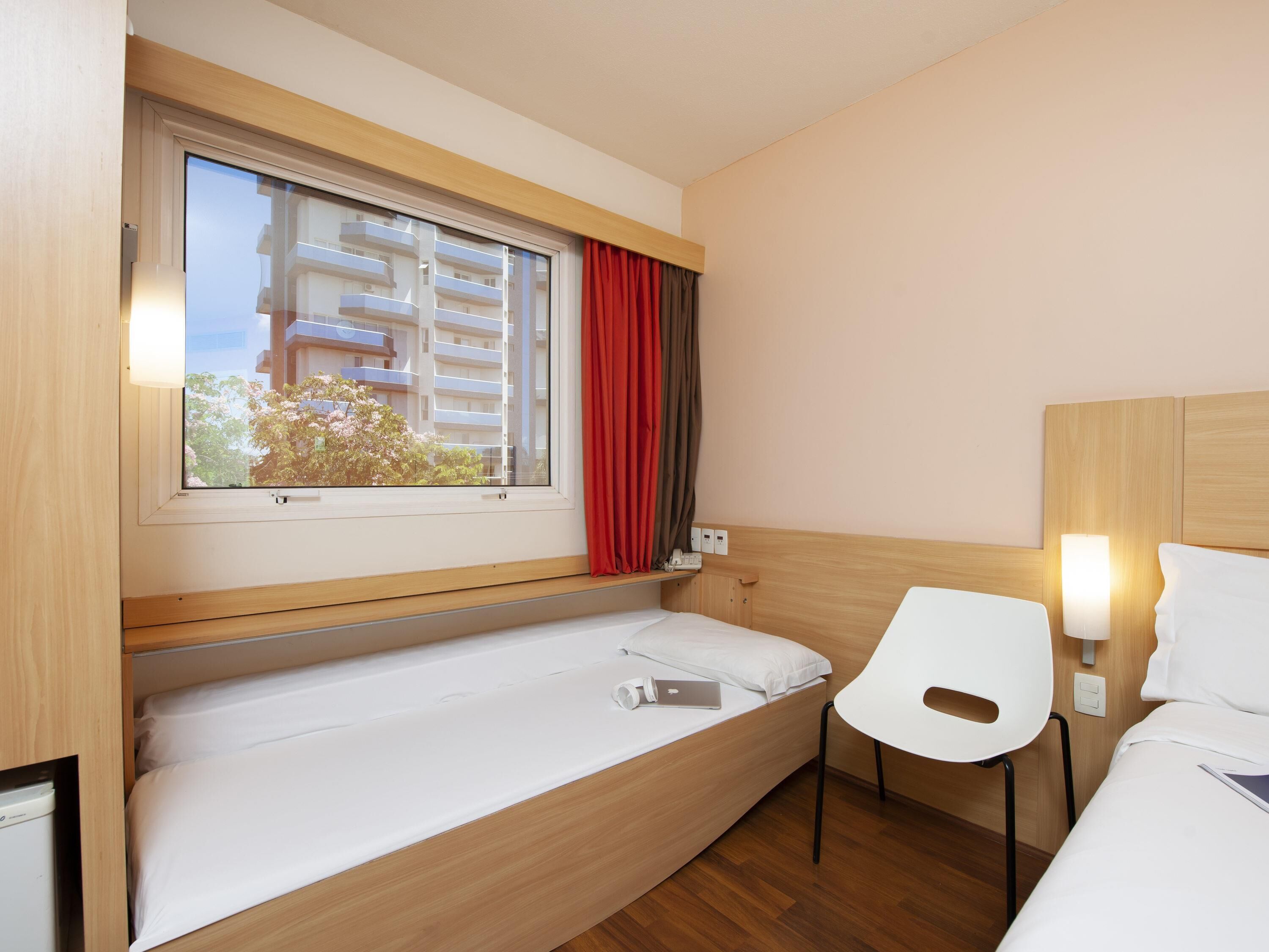 Quarto de hotel compacto com uma cama de solteiro, uma pequena escrivaninha, uma cadeira, uma janela com cortinas e uma luminária fixada na parede.