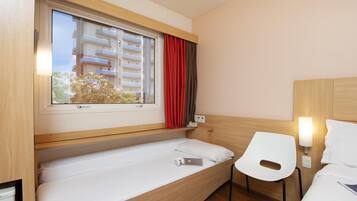 Quarto de hotel compacto com uma cama de solteiro, uma pequena escrivaninha, uma cadeira, uma janela com cortinas e uma luminária fixada na parede.