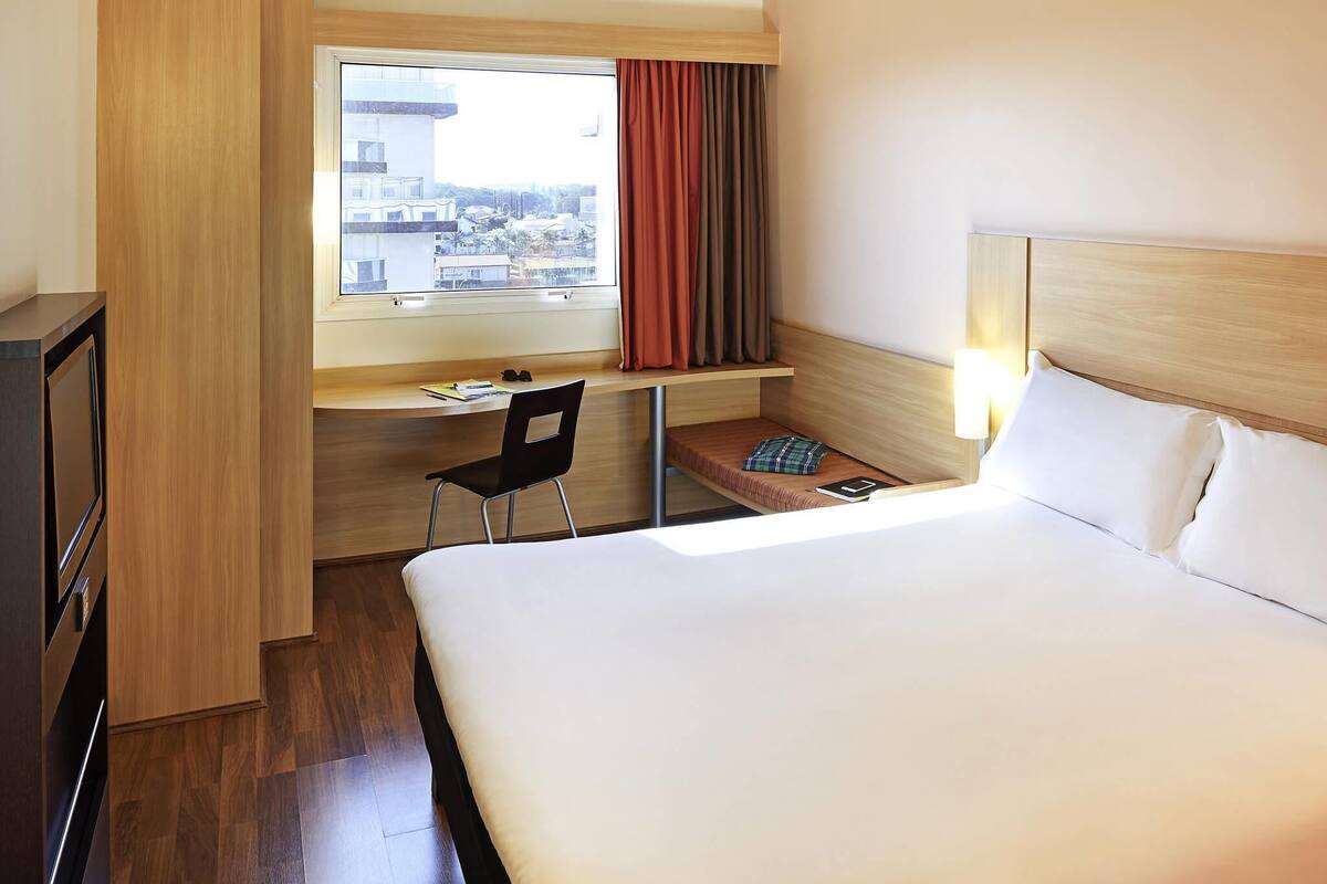 A hotel room with a bed, a desk with a chair, a window with curtains, and a city view.