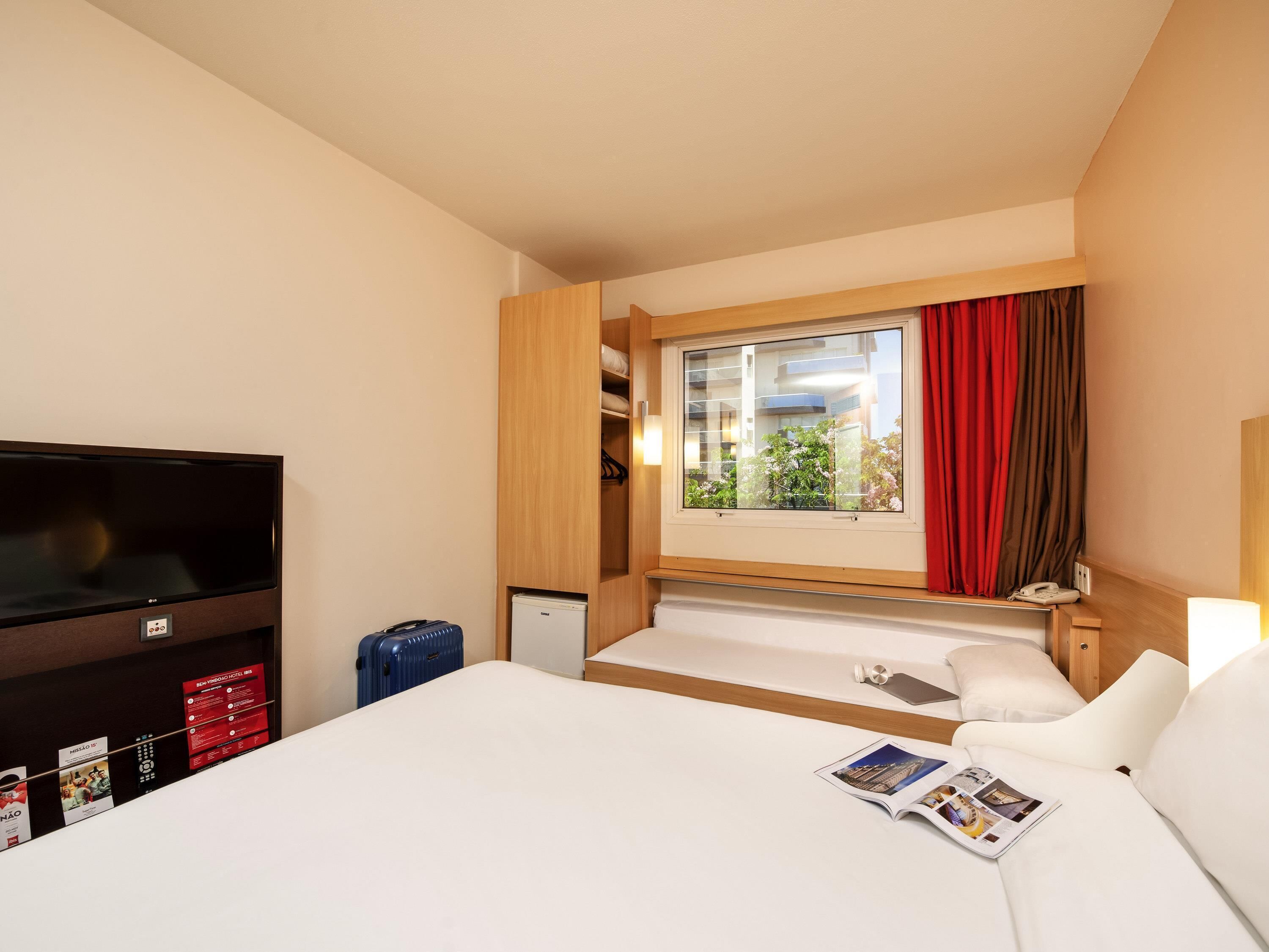 Una habitación de hotel con cama, televisor, ventana con cortinas, una mesita pequeña y un teléfono en la pared.