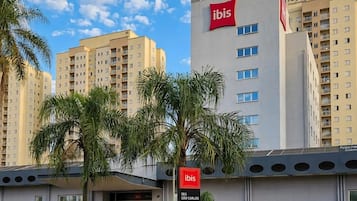 A modern hotel with a prominent ibis logo, surrounded by palm trees and under a clear sky.