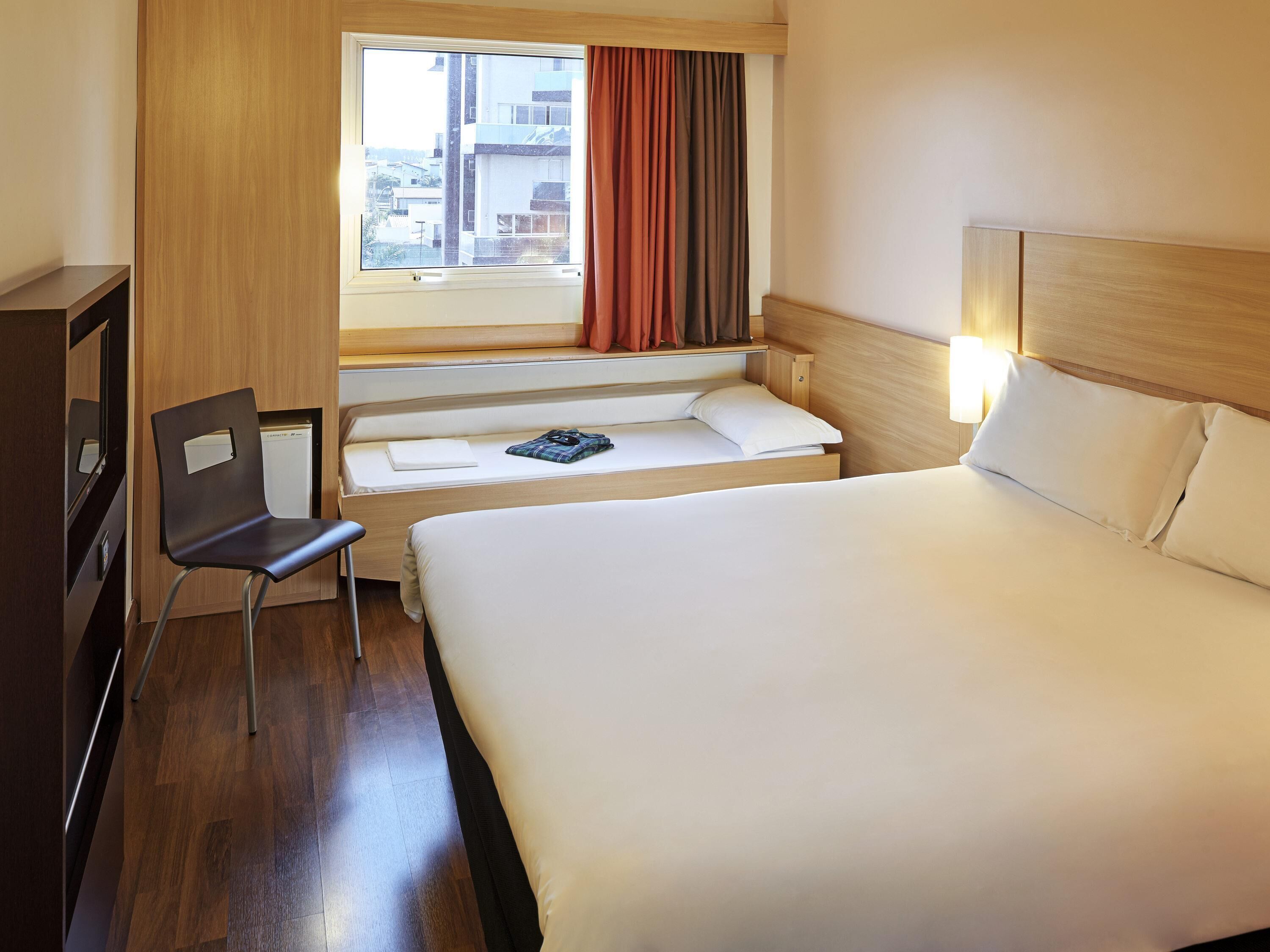 Ein Hotelzimmer mit Bett, Schreibtisch, Stuhl, einem Fenster mit Vorhängen und Blick auf Gebäude.
