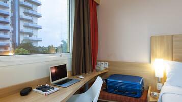 A hotel room with a wooden desk, a laptop, a chair, a bed with a white comforter, a blue suitcase, and a window with curtains.