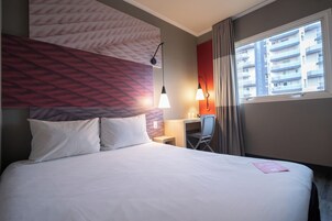Habitación de hotel con cama, escritorio, silla y ventana con vistas a edificios.