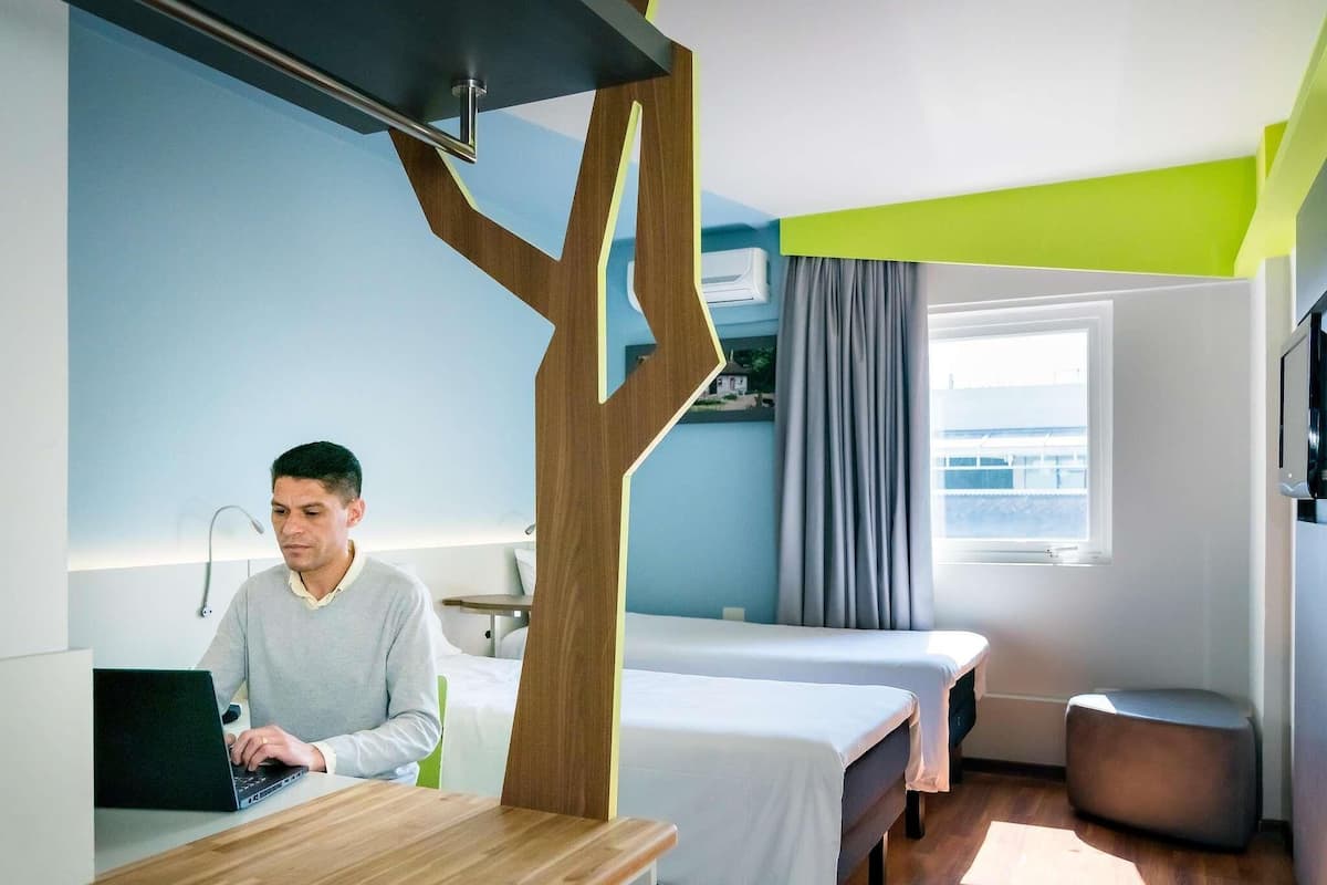 Une chambre d’hôtel avec un mur d’accent en bois, un bureau avec un ordinateur portable, deux lits, une fenêtre avec des rideaux et un téléviseur fixé au mur.