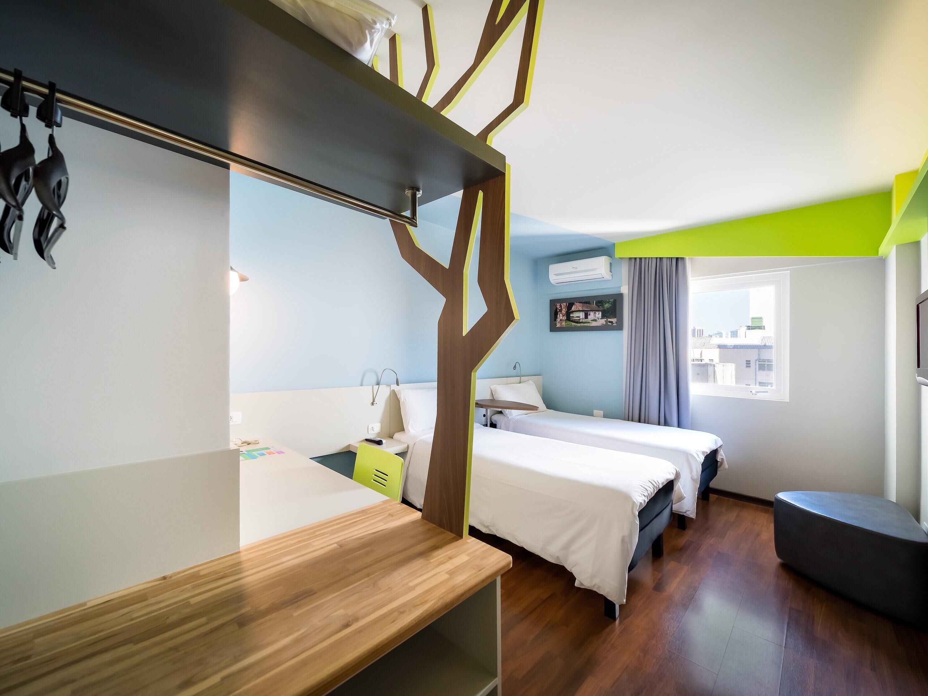 Habitación de hotel con dos camas, un escritorio, una silla, un televisor y ventana con vista a la ciudad.
