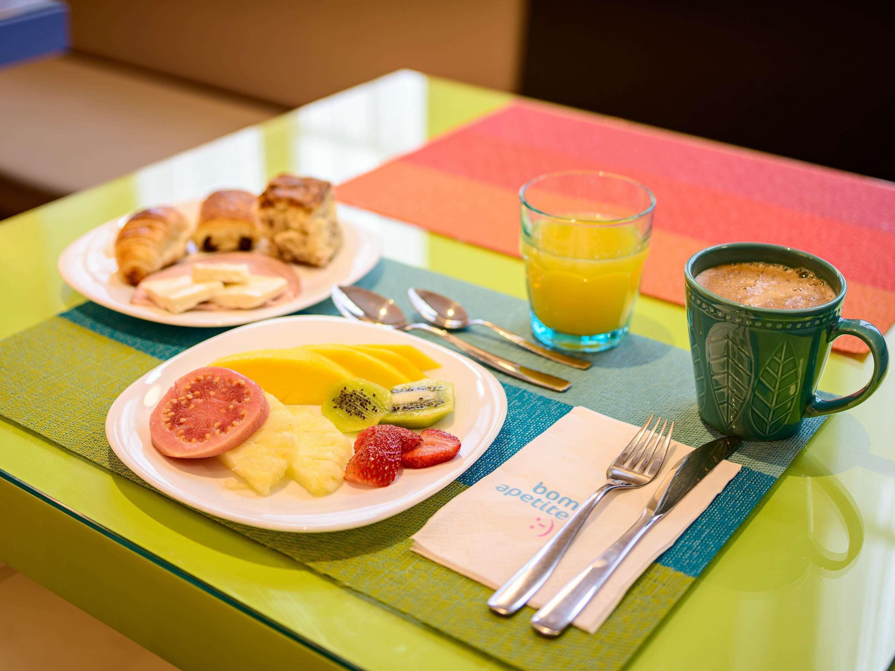 Mesa posta para o café da manhã com uma travessa de frutas, croissants e uma xícara de café.