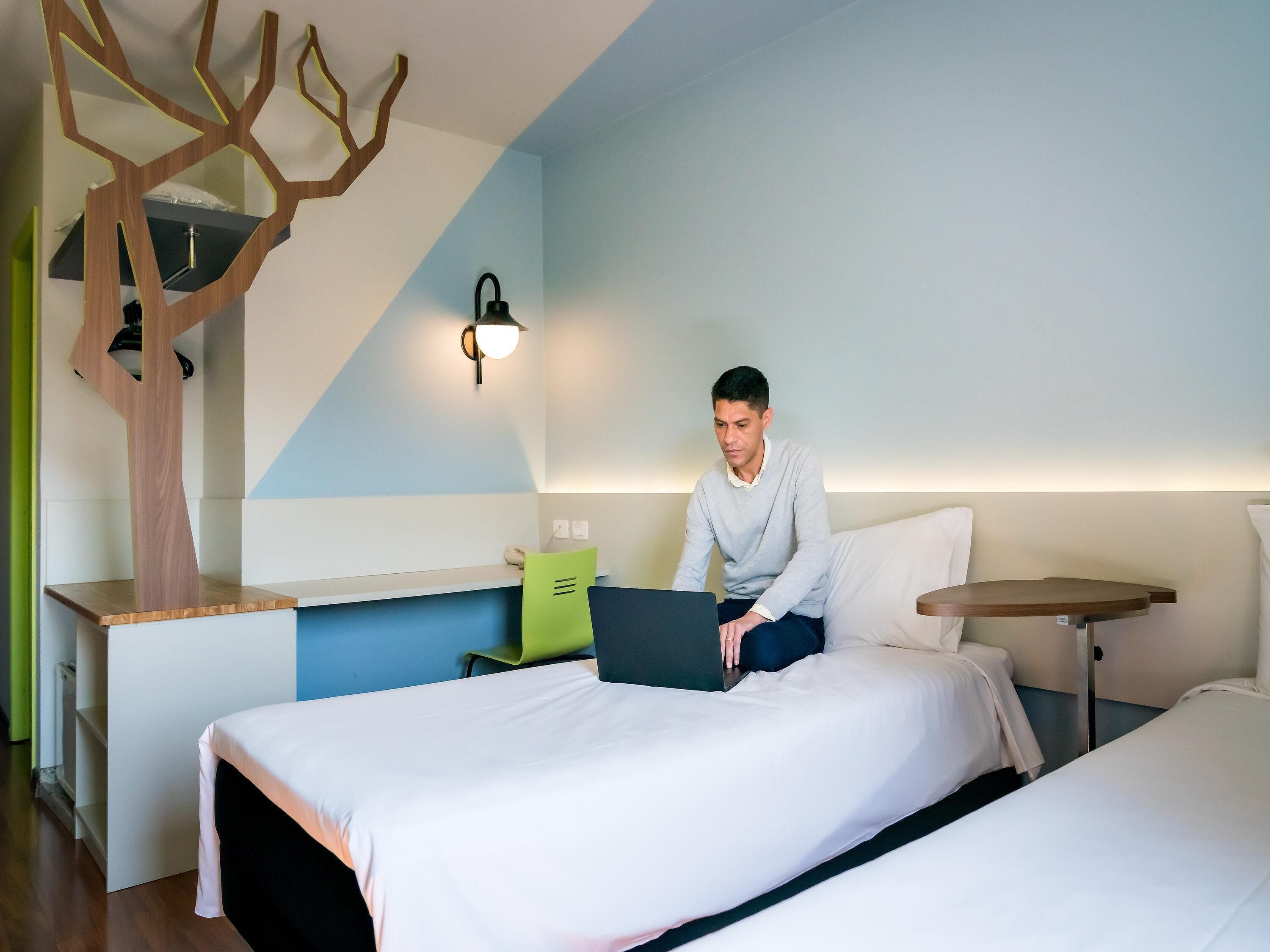 Habitación de hotel con cama, escritorio con una computadora portátil, una mesita y una lámpara fijada a la pared.