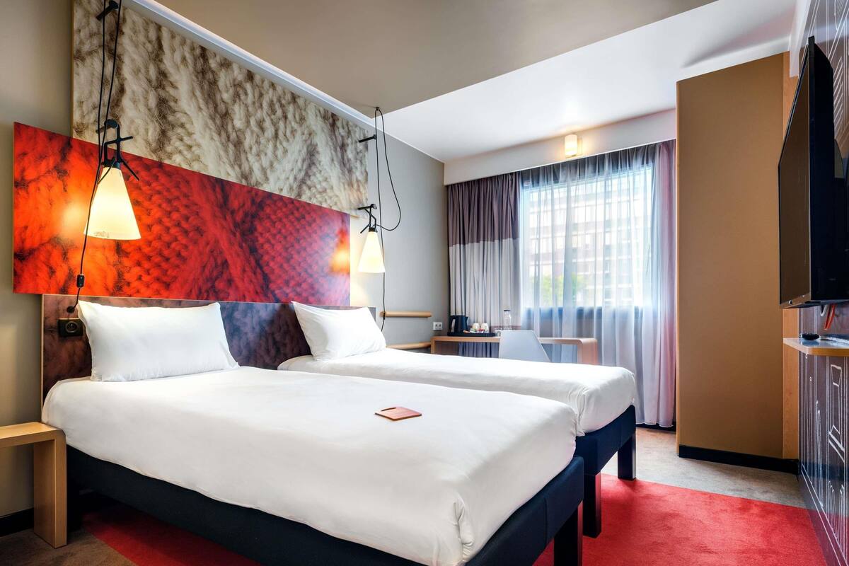 Une chambre d’hôtel avec deux lits, un bureau, une télévision et une grande fenêtre avec des rideaux.