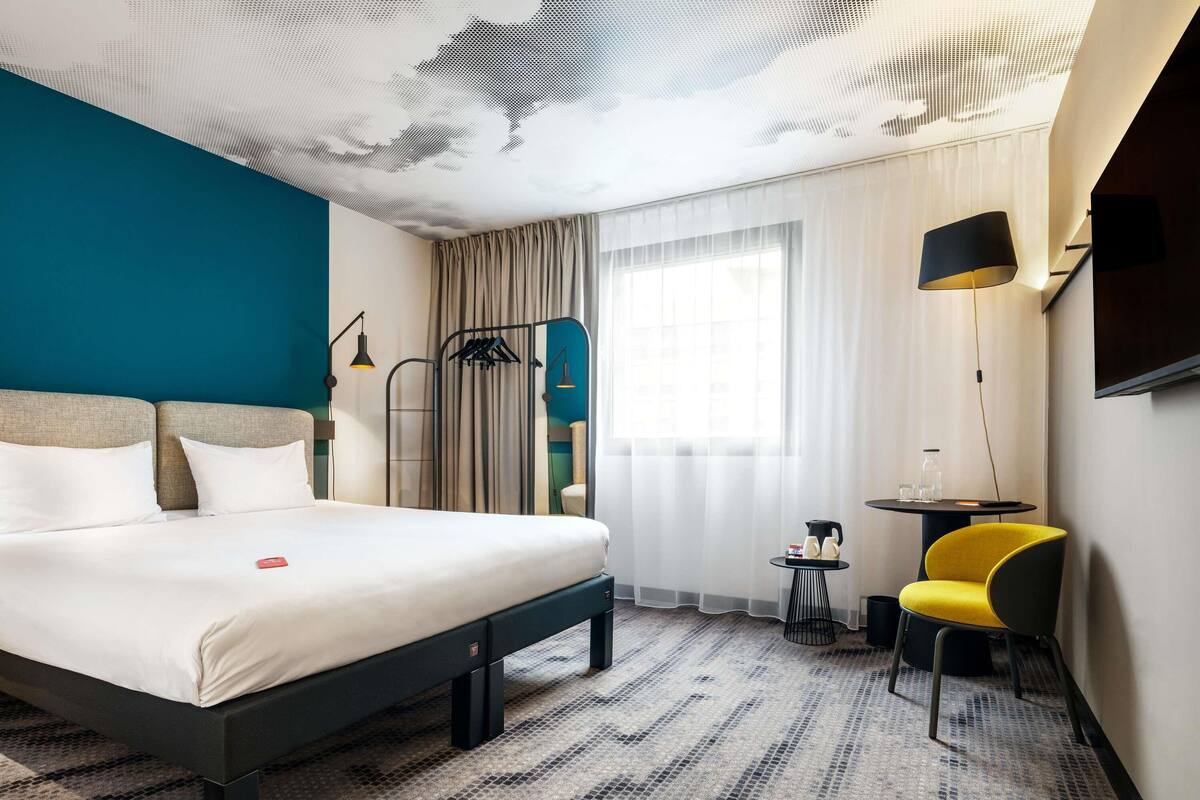 Une chambre d’hôtel moderne, dotée d’un grand lit, d’une petite table avec une chaise, d’une télévision et d’une fenêtre avec des rideaux.