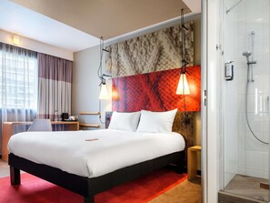 Ett hotellrum med en stor säng, ett skrivbord, en stol, en dusch och utsikt över staden.