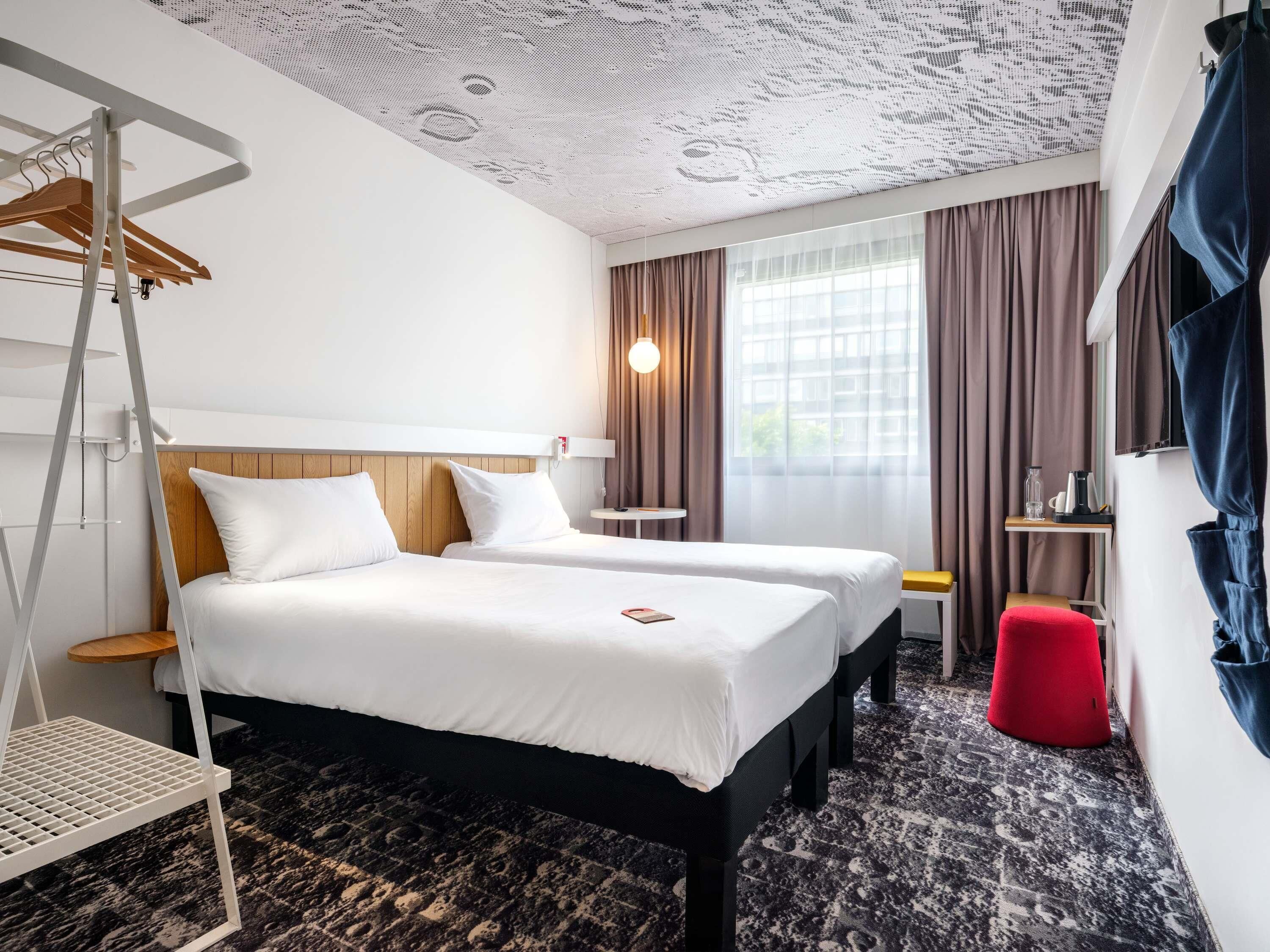 Hotelkamer met twee bedden, een rode poef, een klein tafeltje met lamp en een plafond met een uniek patroon.