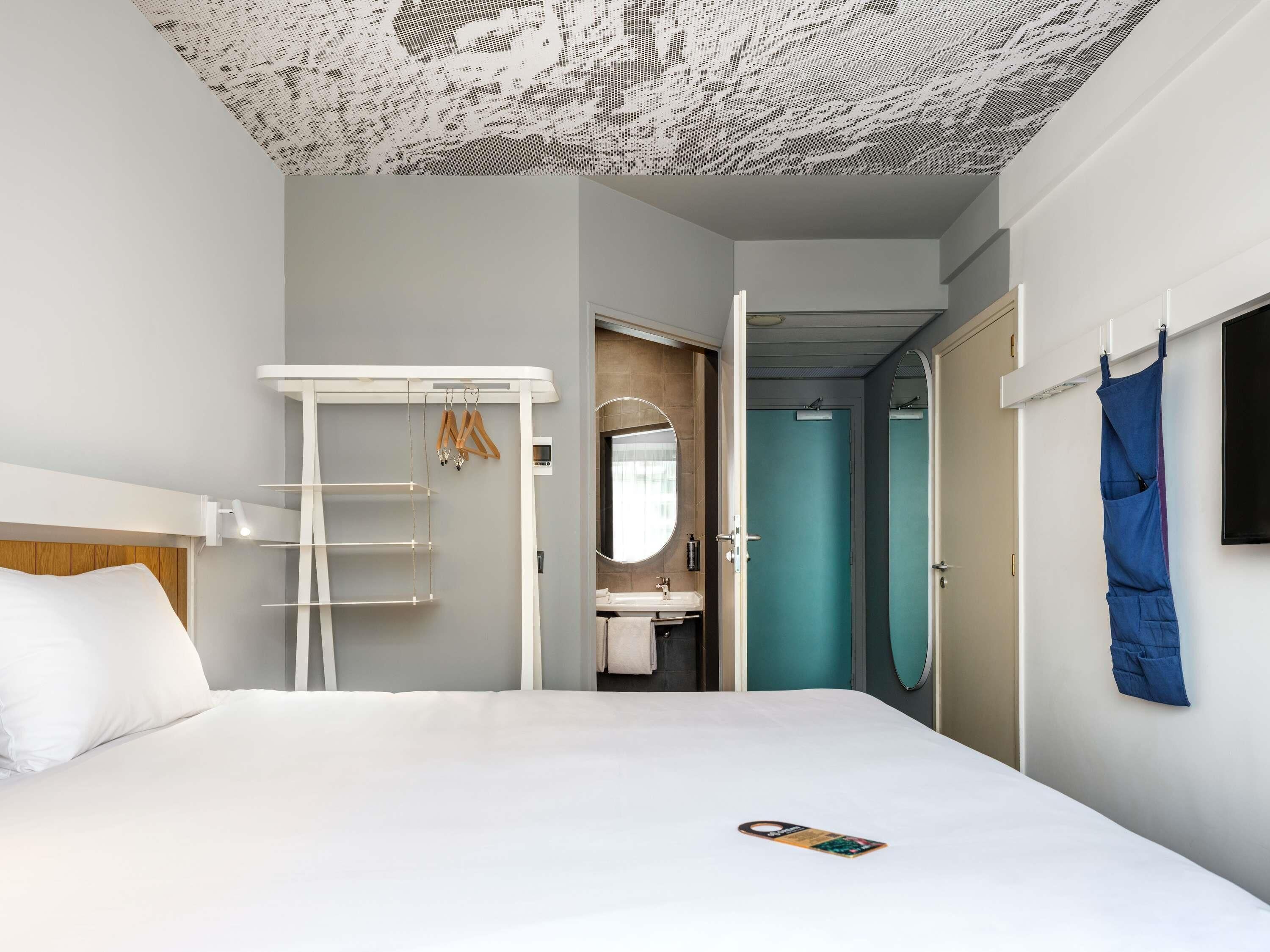 Une chambre d’hôtel moderne avec un lit, une armoire blanche, une salle de bain avec un lavabo et un miroir, et un vêtement bleu accroché au mur.