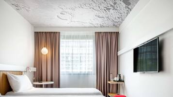 A hotel room with a bed, a TV, a small table with a lamp, and a window with curtains.
