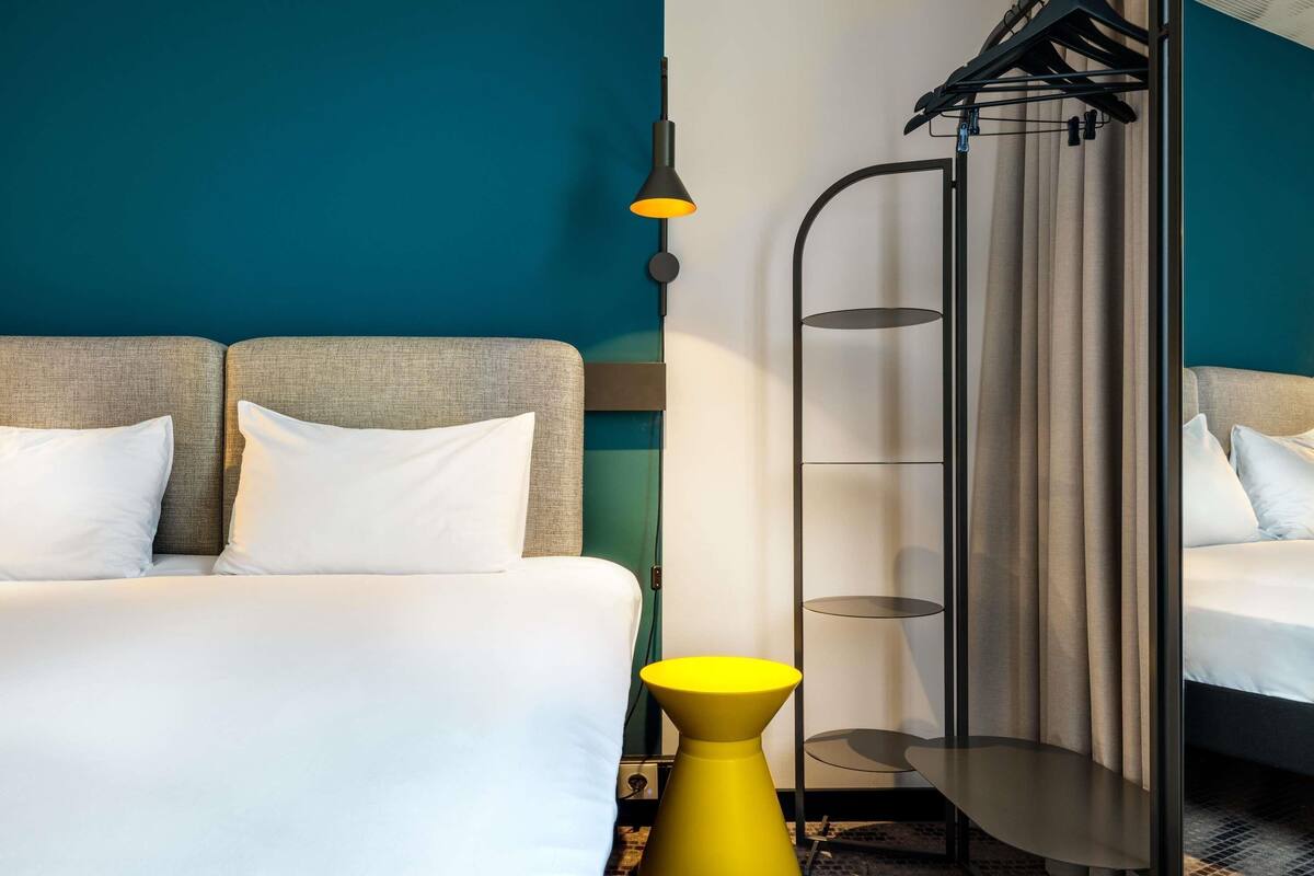 A hotel room with a bed, a yellow side table, a black hanging rack, and a floor lamp.