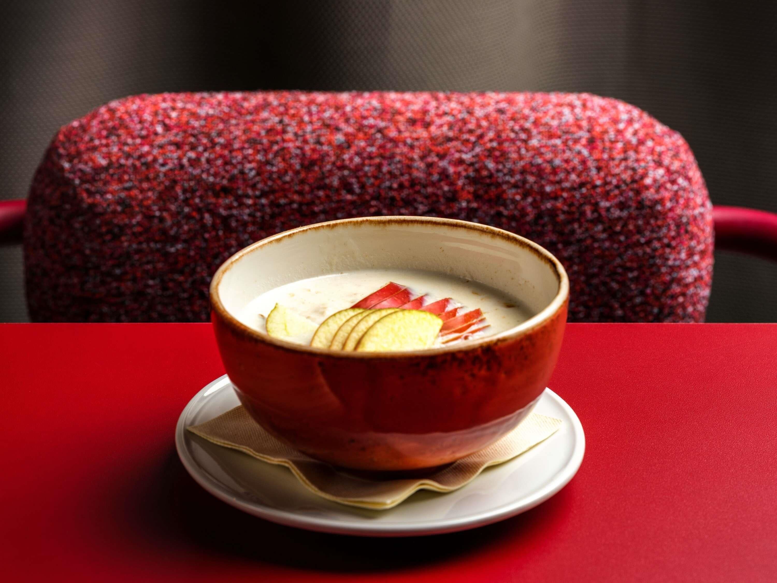 A bowl of soup with apple slices on a white plate, placed on a red table with a red chair in the background.