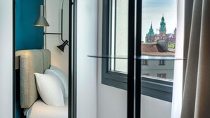 A modern hotel room with a bed, a yellow stool, a small table, and a view of a historic building through a window. - ibis Krakow Centrum (Kraków)