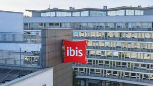 A modern building with a prominent red 'ibis' sign, multiple windows, and a parking area with a street view. - ibis Krakow Centrum (Kraków)