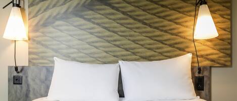 Una habitación de hotel con una cama, dos lámparas fijadas a la pared y un revestimiento texturizado detrás de la cama.