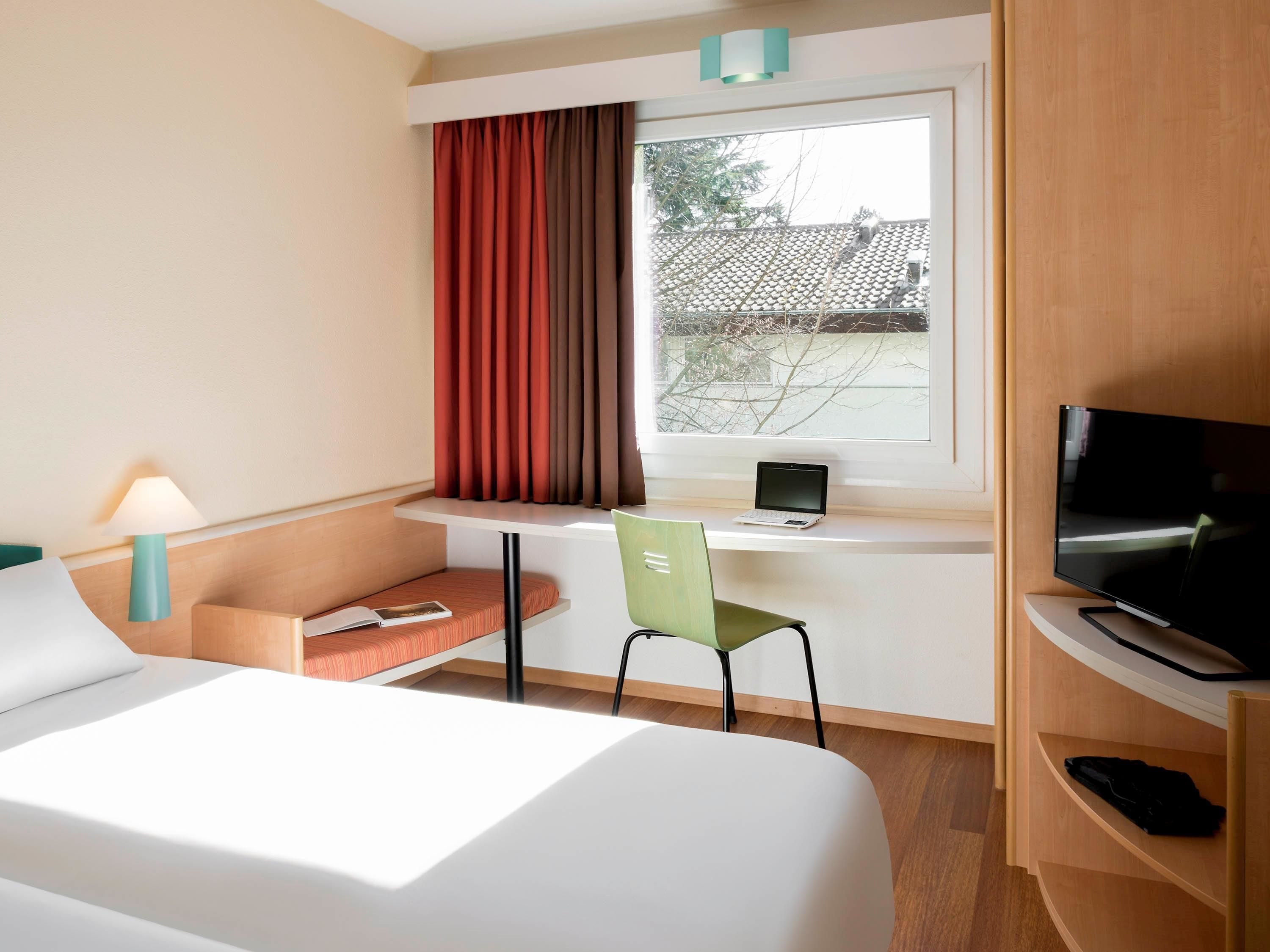 A hotel room with a bed, a desk with a laptop, a TV, a chair, and a lamp.
