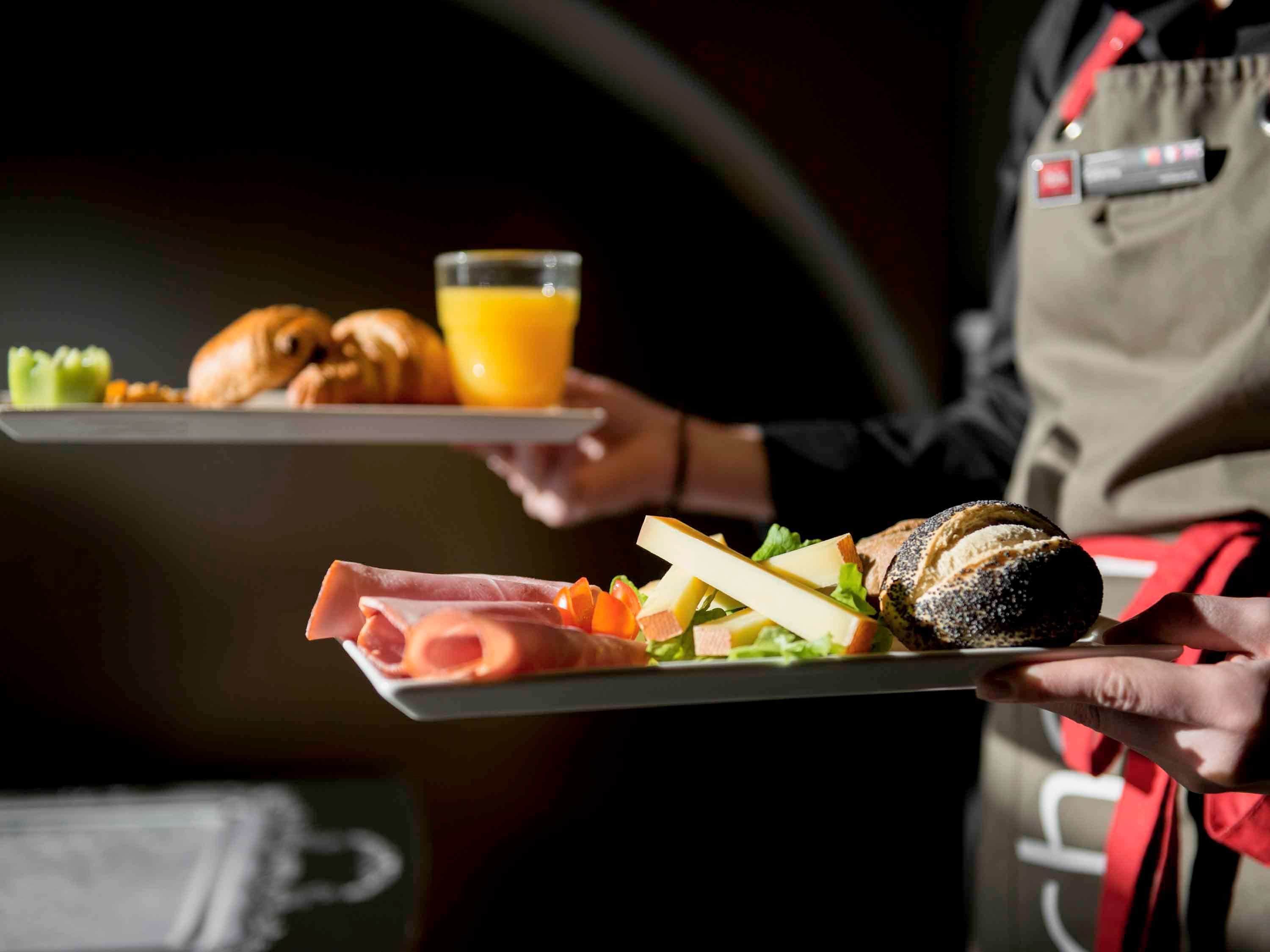 Eine Person hält ein Tablett mit Speisen, darunter Croissants, ein Glas Orangensaft und ein Sandwich mit Käse und Gemüse.