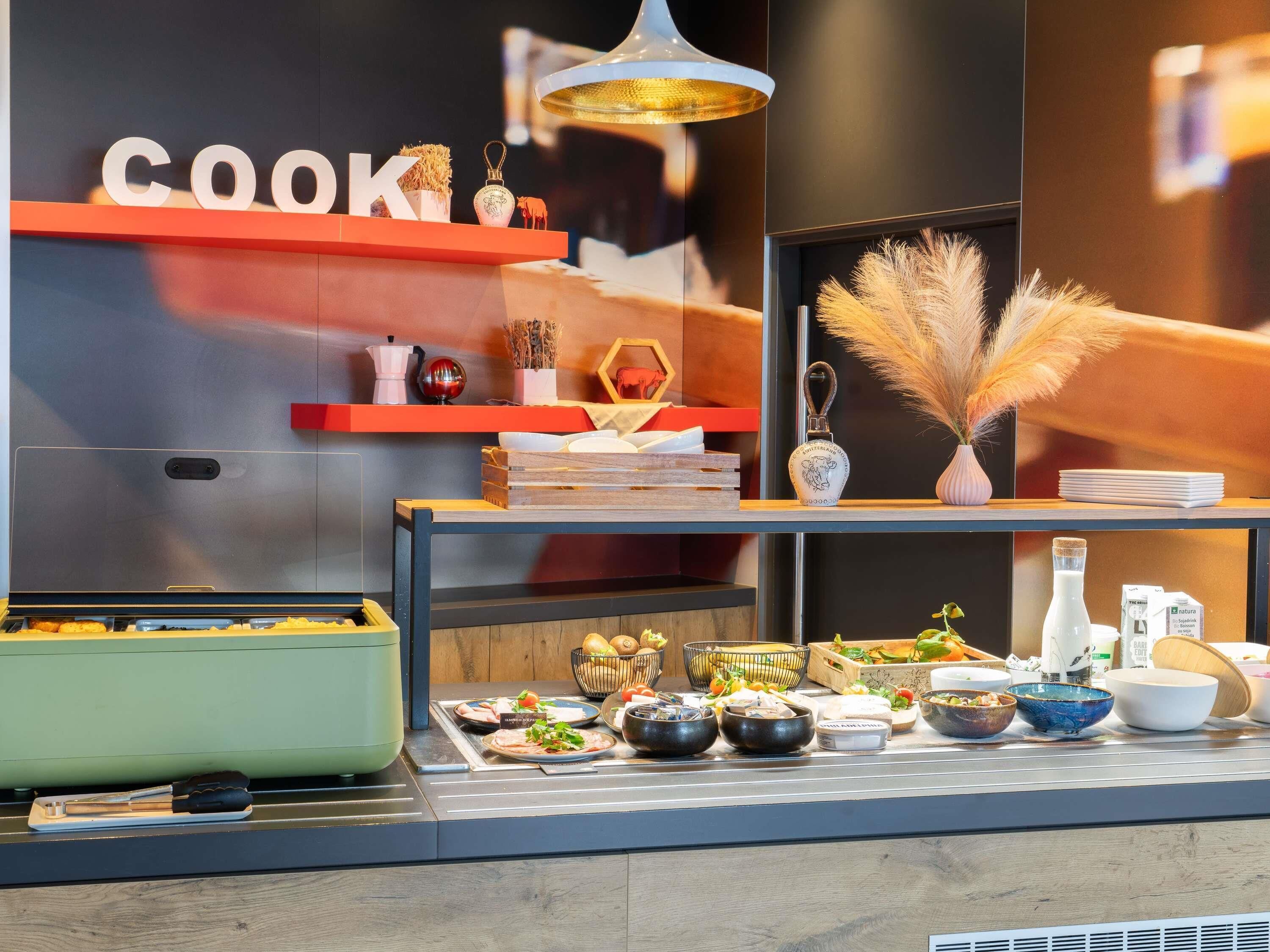 Una zona de cocina con una encimera que exhibe diferentes platos y utensilios, un estante con adornos y una pared donde se lee la palabra 'COOK' en grande.
