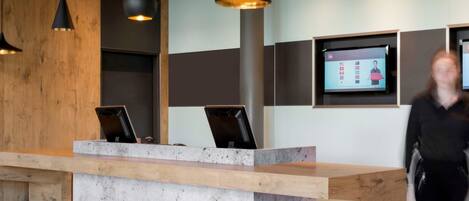 A reception desk with a modern design, featuring a concrete base and wooden accents. There are two black pendant lights above the desk, and a flat-screen TV mounted on the wall to the right.