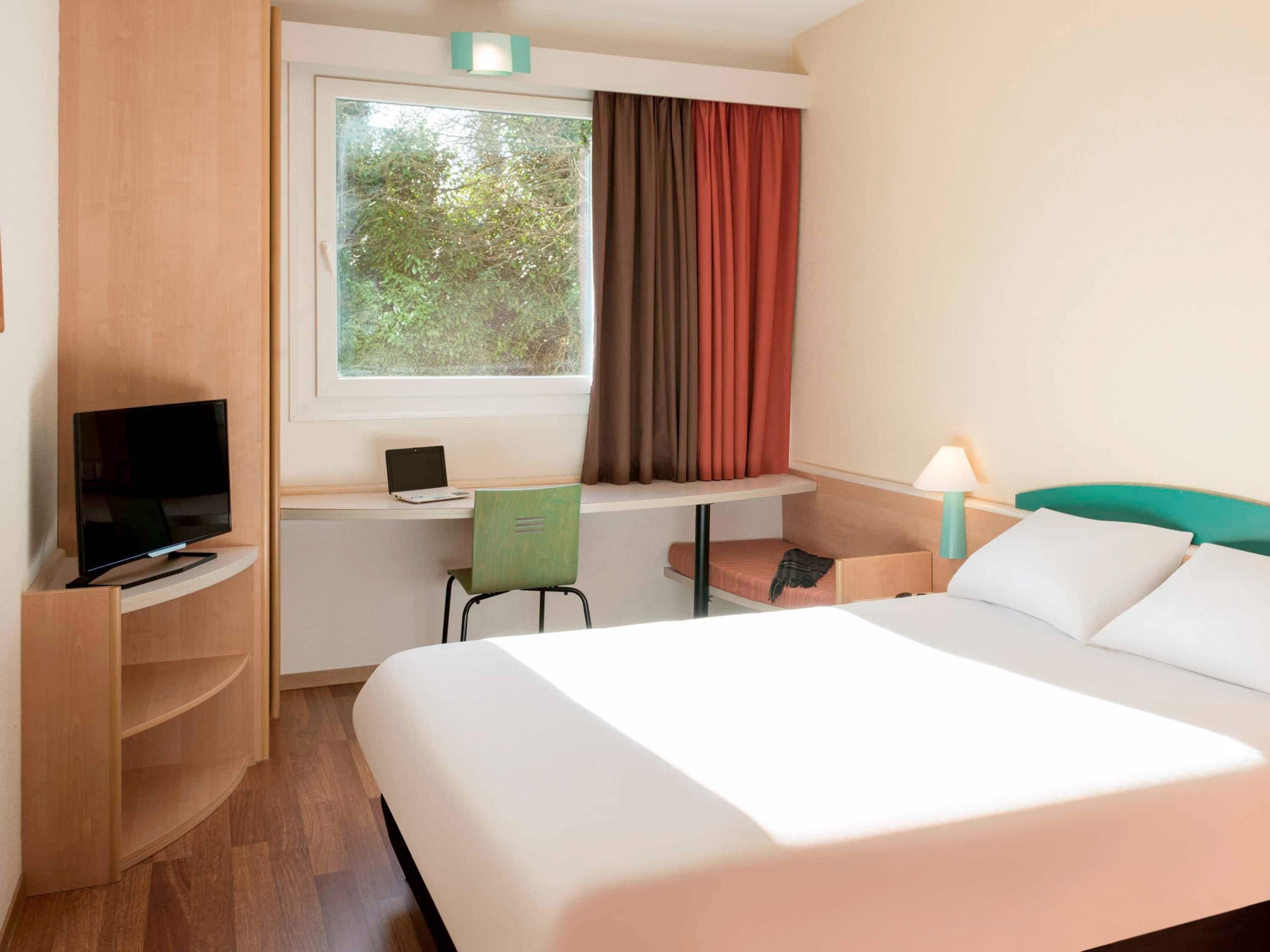 Ein Hotelzimmer mit Bett, Schreibtisch mit Laptop, Fernseher, Stuhl und einem Fenster mit Vorhängen.