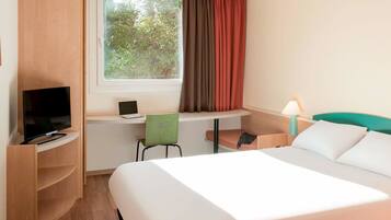 Ein Hotelzimmer mit Bett, Schreibtisch mit Laptop, Fernseher, Stuhl und einem Fenster mit Vorhängen.