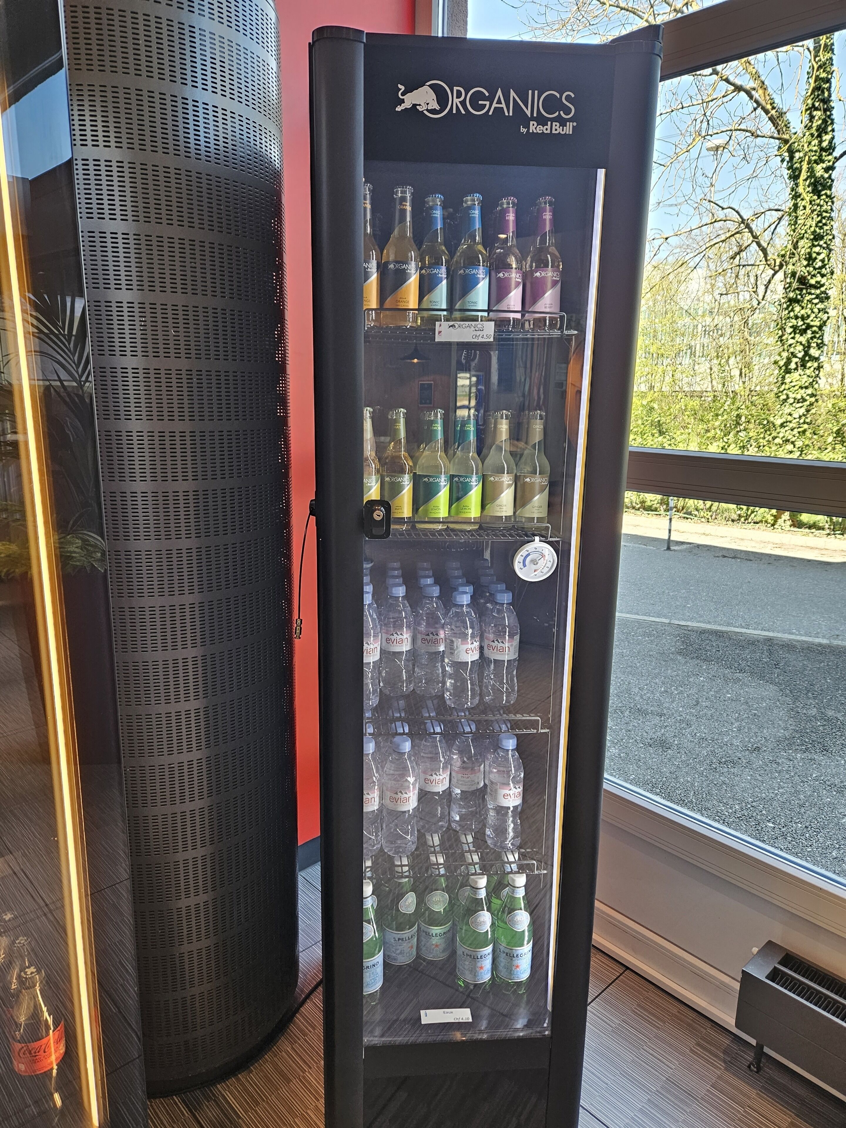 A beverage vending machine with multiple drink options, including water and various alcoholic beverages, located indoors near a window.