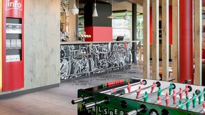 A modern indoor space with a foosball table, a red information stand, and a bar area with a black and white mural. - ibis Geneve Aeroport (Meyrin)