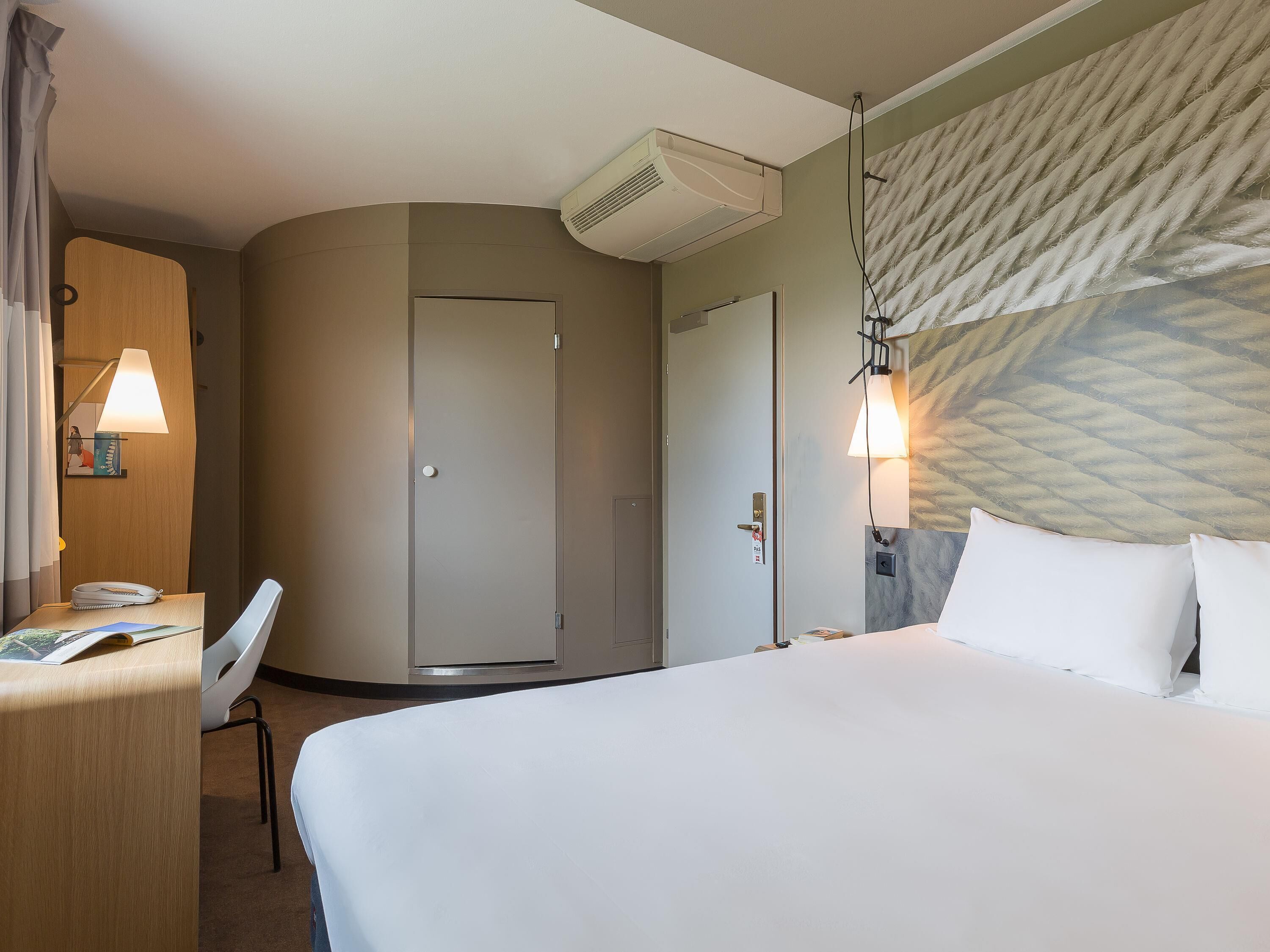Quarto de hotel com cama, escrivaninha, cadeira, ar condicionado e luminária na parede.
