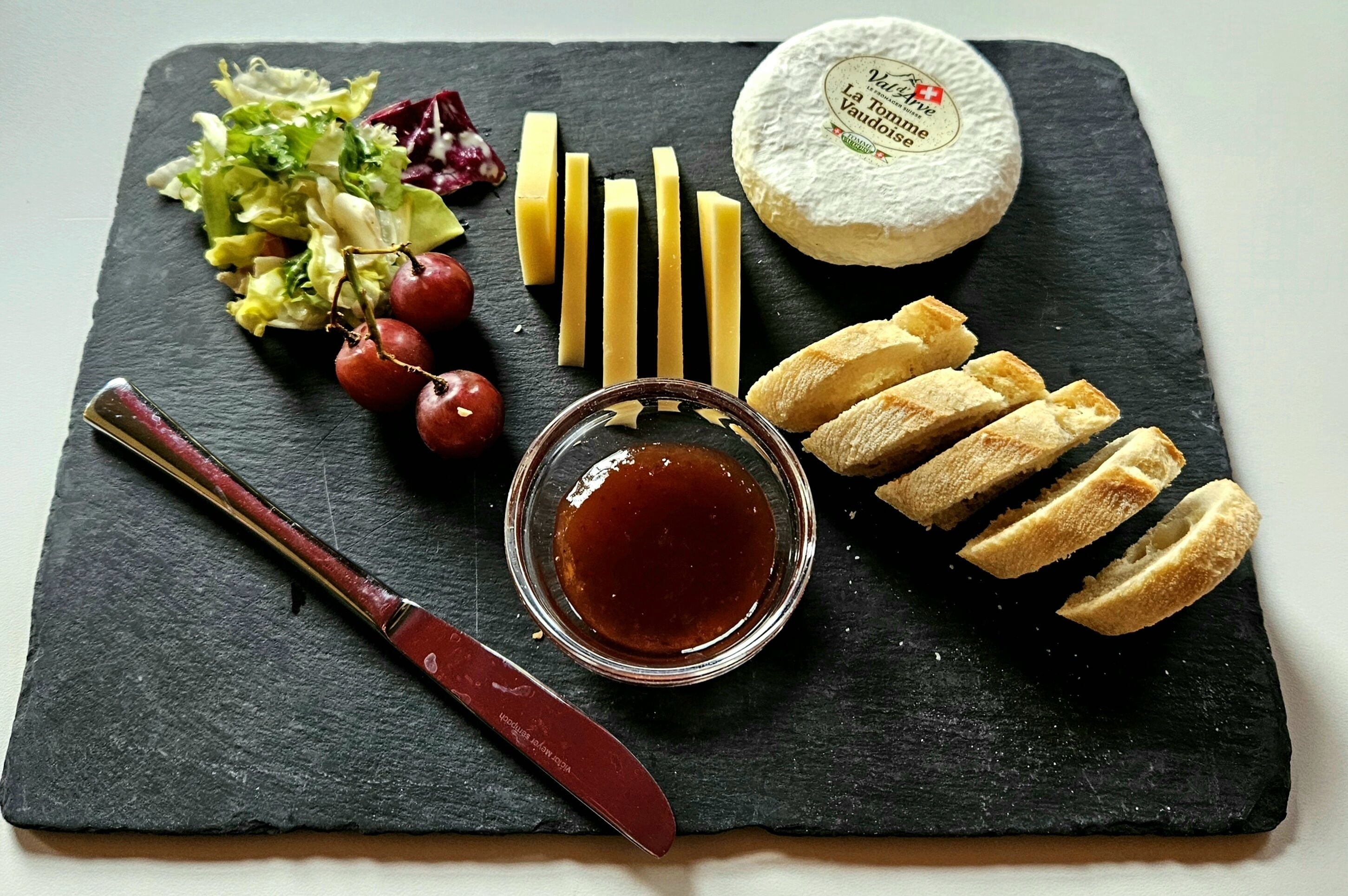 Une planche de fromages comprenant différents types de fromages, des raisins et du pain, le tout présenté sur une ardoise.