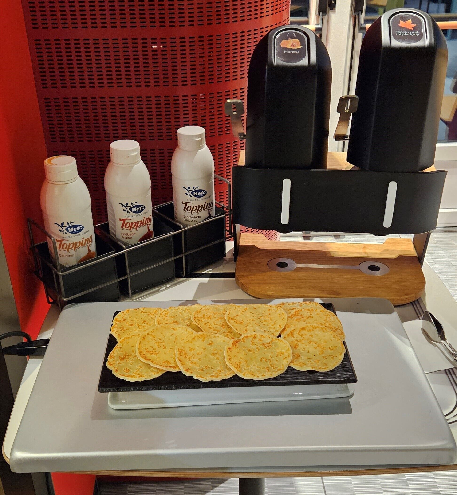 A table with a stack of golden-brown pancakes and three bottles of Hero Topping Caramel, Chocolate, and Chocolate Sauce.