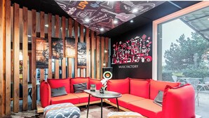Un rincón tranquilo con un sofá rojo, otomanas estampadas y una mesita. Se aprecia un mural en la pared con instrumentos musicales y la inscripción "MUSIC FACTORY".