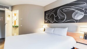 Habitación de hotel con una cama grande, una mesita de noche, un cuadro colgado en la pared y un baño visible a través de una puerta abierta.