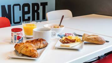 A breakfast table with croissants, scrambled eggs, bacon, toast, a glass of orange juice, a coffee cup, and a bowl of cereal.