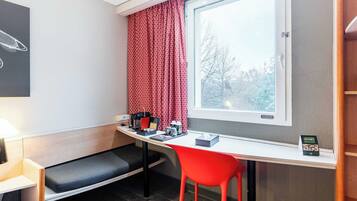 Ett litet rum med ett skrivbord, en röd stol, en bänk, ett fönster med gardiner och ett litet bord med en lampa och en skylt.