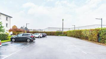 A parking lot with several cars, a building in the background, and a hedge along the side.