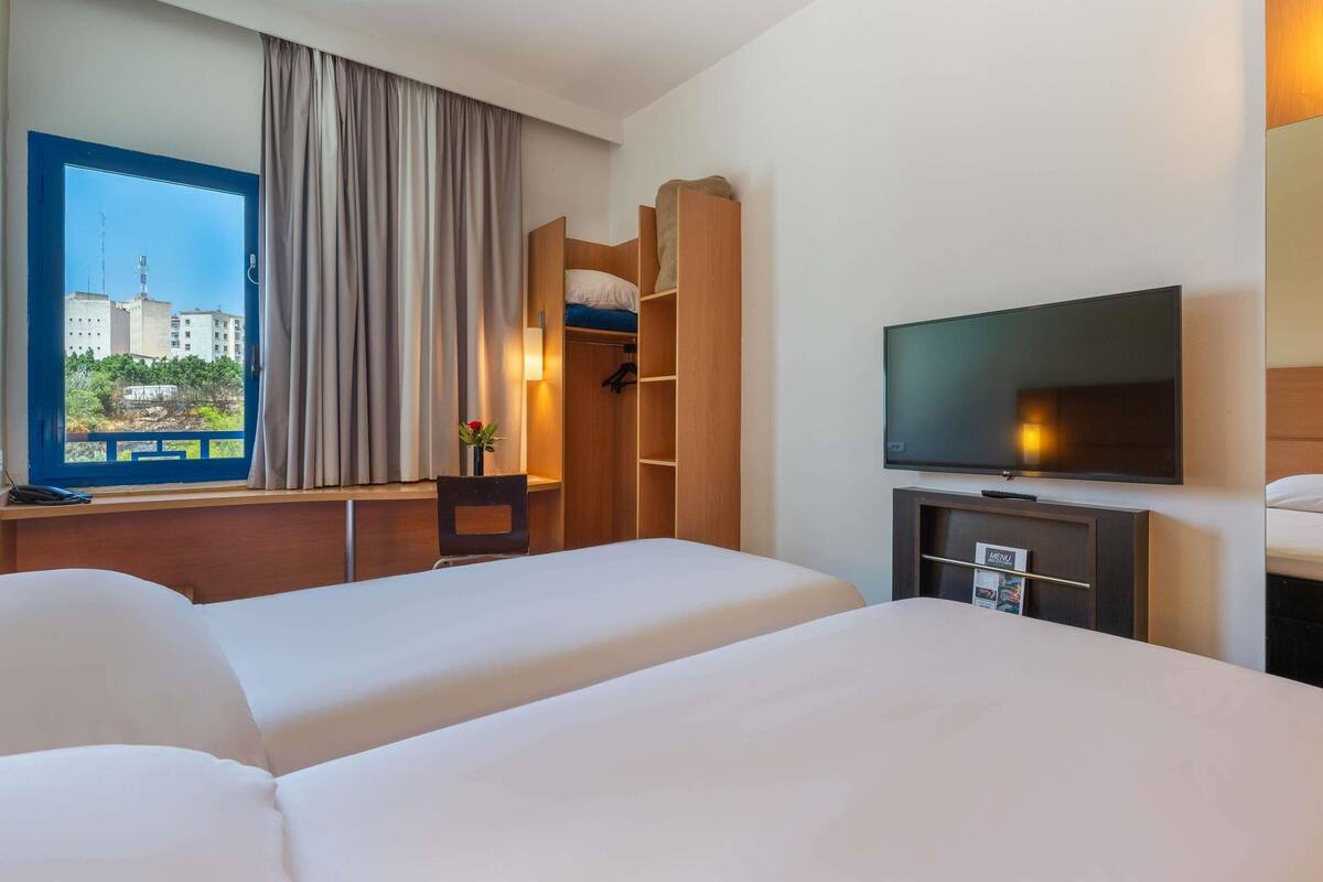 A hotel room with two beds, a TV, a desk with a lamp, and a window with curtains.