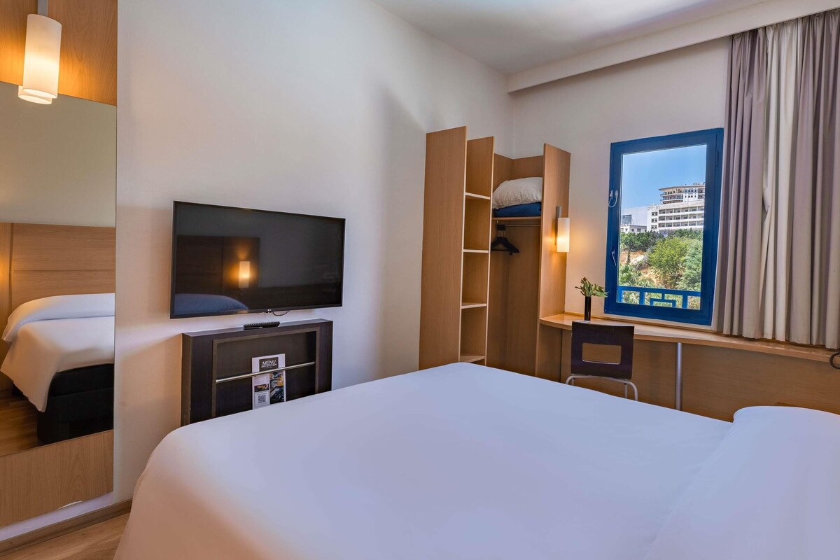 A hotel room with a bed, a TV, a desk, and a window with a view of buildings.