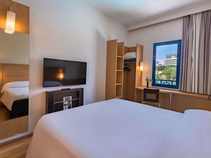 A hotel room with a bed, a TV, a desk, and a window with a view of buildings. - ibis Meknes Hotel (Meknes)